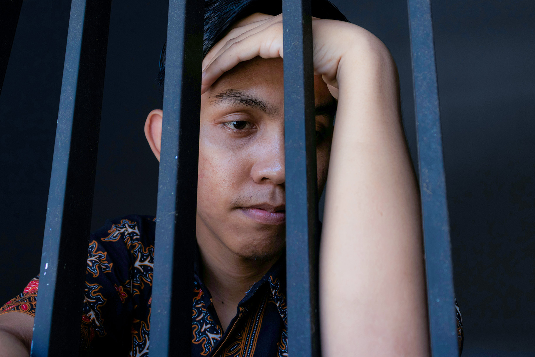 Imagen de un joven hispano sujetándose la cabeza con incredulidad, de pie tras las rejas en un centro de detención.