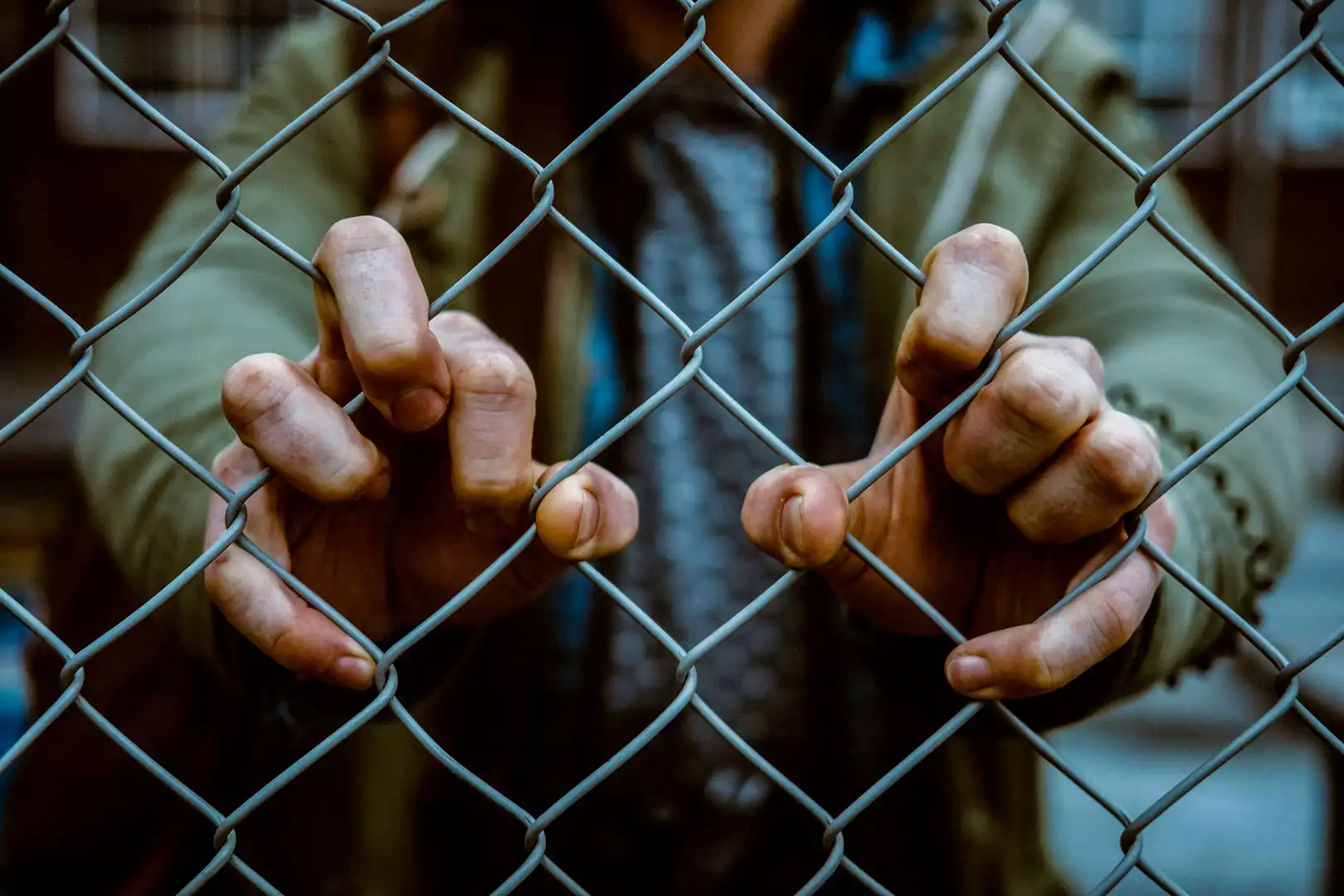 Image of a man clenching a fence with his fingers, appears to be confined in a US detention facility.