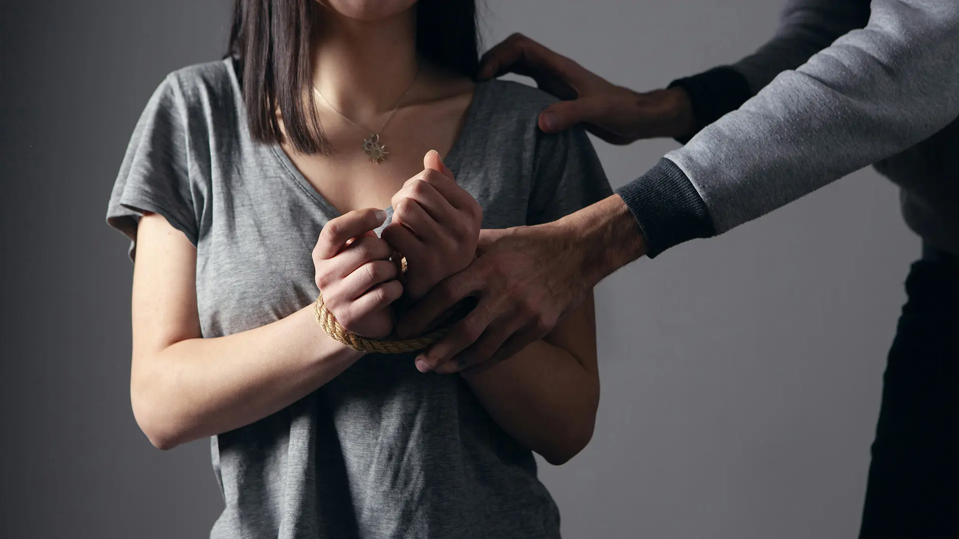Photograph of a woman crouched down with her hands bound and a male figure standing above her in a menacing manner.