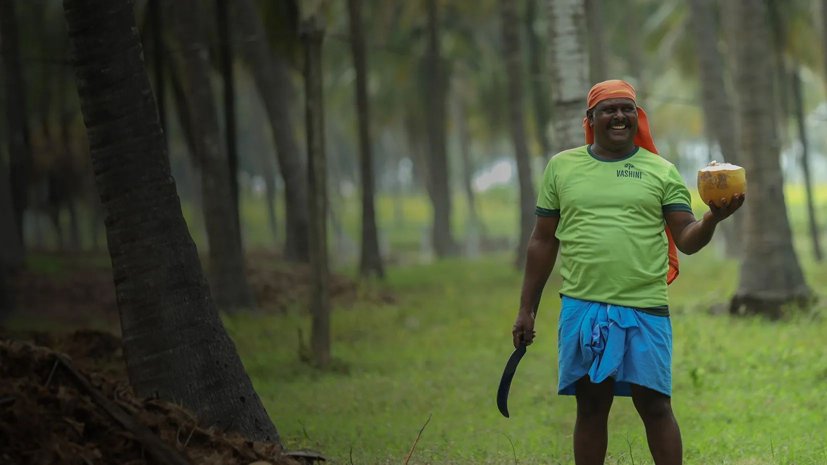 Smiling man in a green shirt and blue wrap holding a cut coconut and a machete standing in a coconut grove.