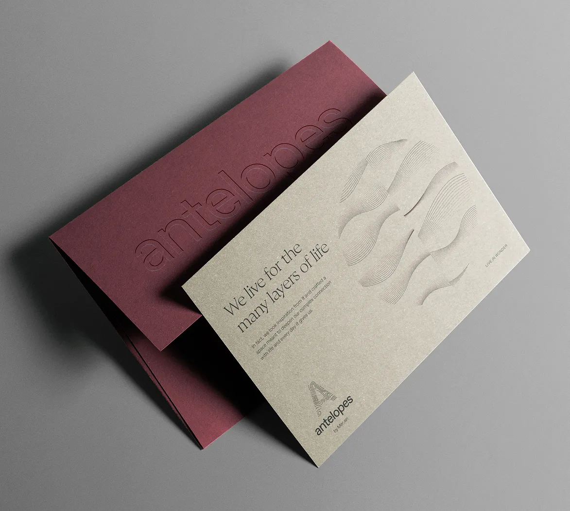 A beige card with wavy line illustrations and text 'We live for the many layers of life' placed on a burgundy envelope embossed with the word 'antelopes'.