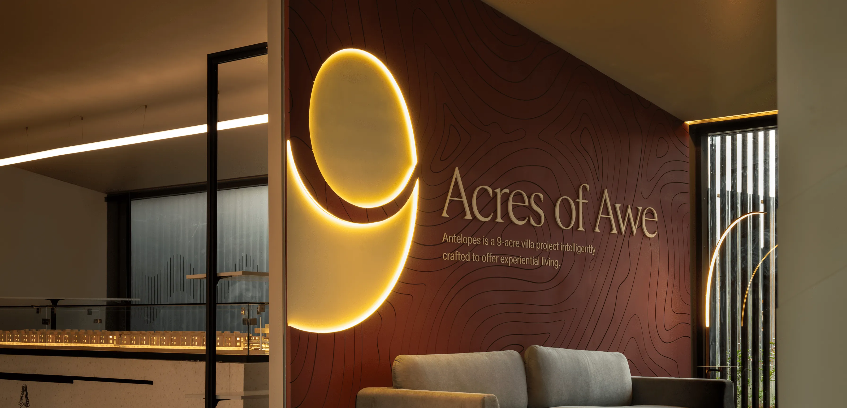 Modern reception area with a lit golden abstract logo and the sign 'Acres of Awe' on a textured dark wall behind a gray couch.