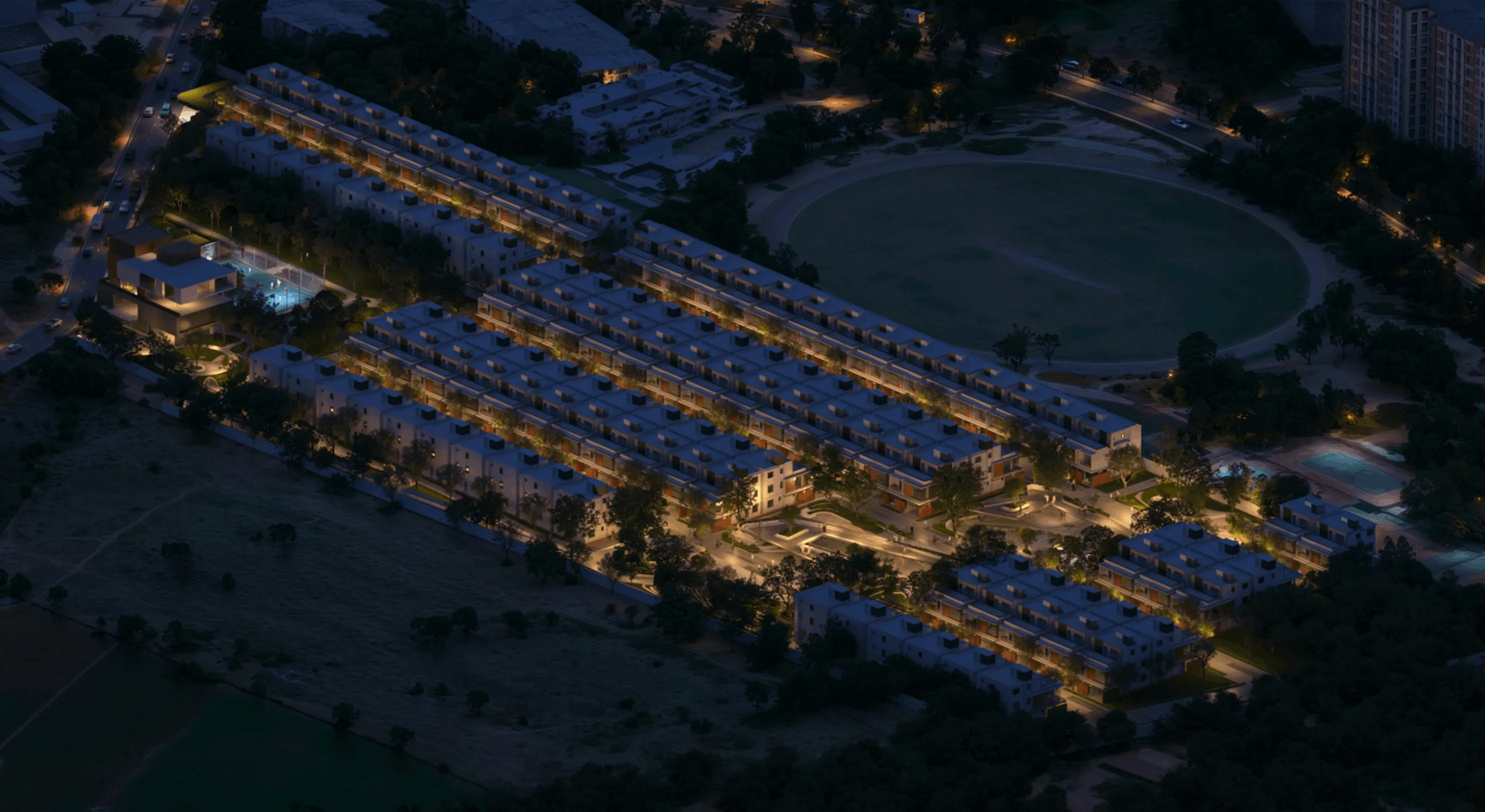 Aerial night view of a modern residential complex with well-lit buildings, roads, trees, and a large circular field nearby.