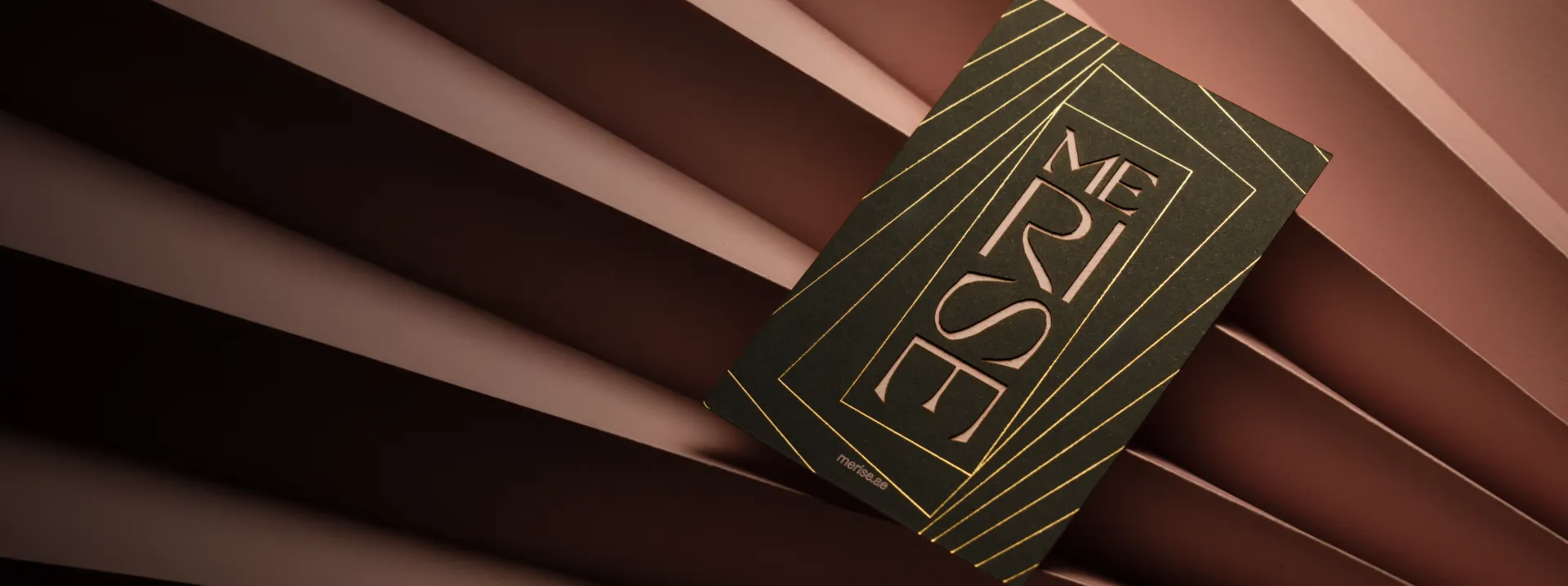 Black card with gold geometric lines and the word 'MERISE' in stylized letters on a brown pleated fabric background.