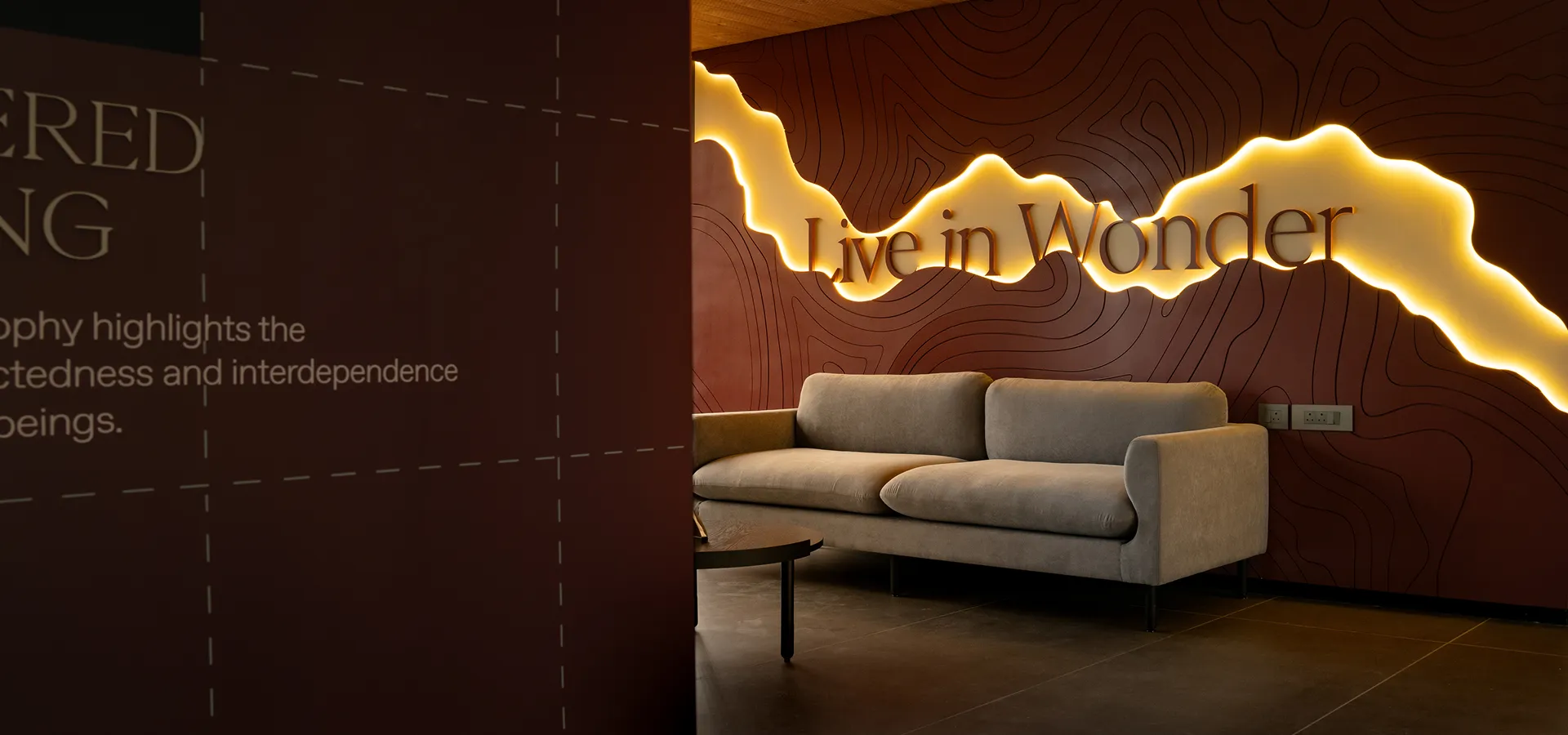 Modern lounge area with a beige sofa under a backlit wall sign reading 'Live in Wonder' on dark brown wall with contour line patterns.