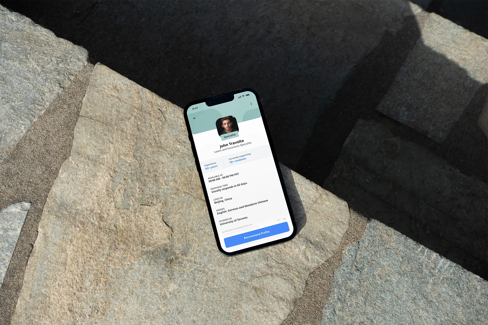 Smartphone on stone pavement displaying a profile of John Travolta, a loans and insurance specialist, with availability, languages, and education details.