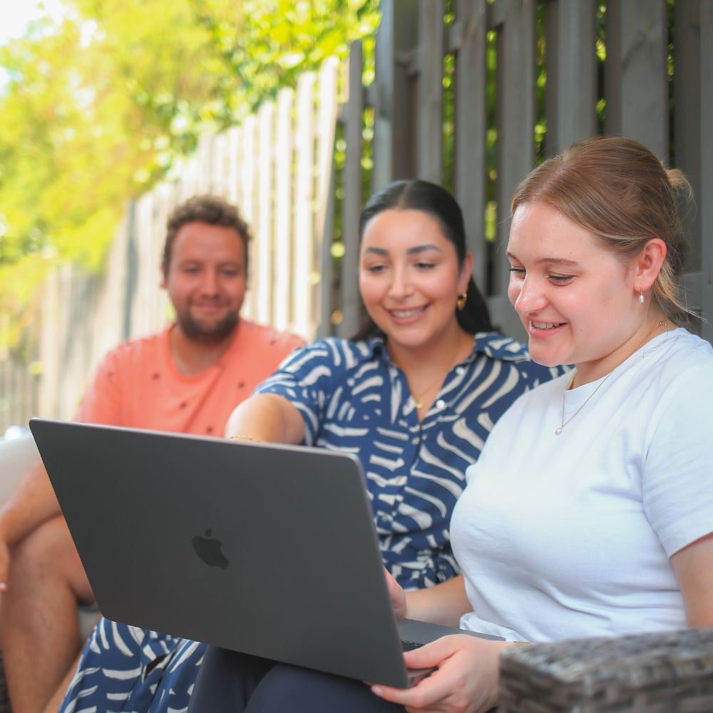 Die Mitarbeiter:innen Janik, Isabella und Jessy sitzen im Außenbereich auf einer Lounge und schauen sich gemeinsam etwas auf dem Laptop an.
