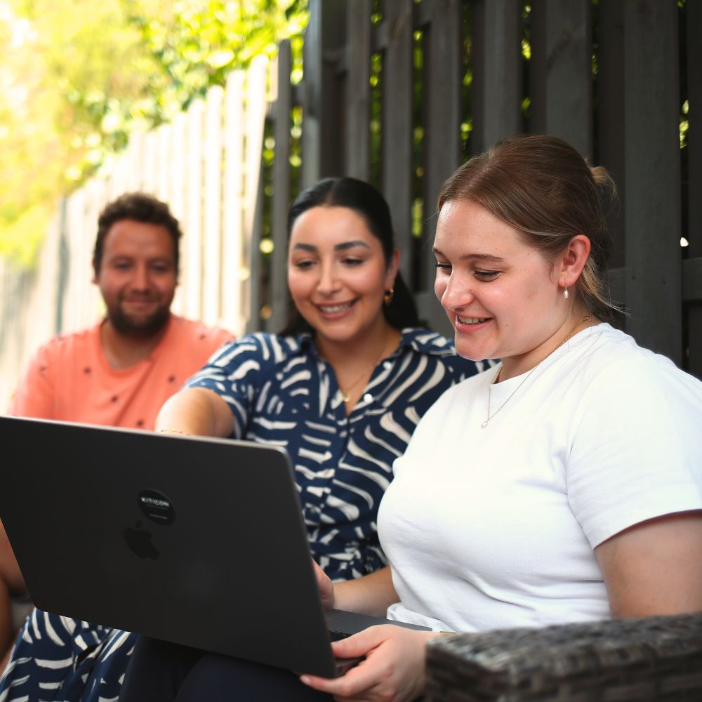 Die Mitarbeiter:innen Janik, Isabella und Jessy sitzen im Außenbereich auf einer Lounge und schauen sich gemeinsam etwas auf dem Laptop an.