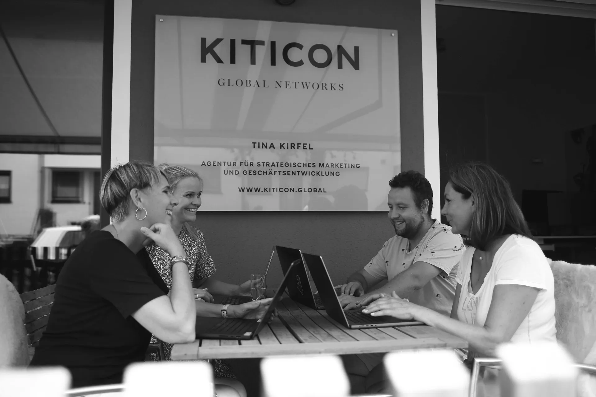 Vier Personen sitzen an einem Tisch mit Laptops und lächeln vor einem Schild mit KITICON-Logo.