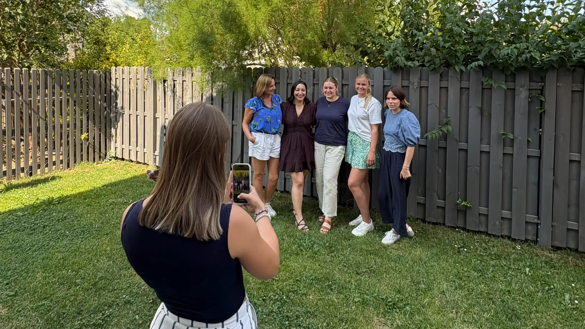 Frau mit schwarzem Oberteil fotografiert Gruppe von fünf Frauen vor einem Holzzaun im Garten