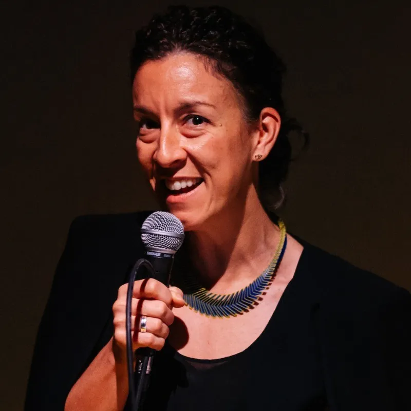 Woman with dark curly hair speaking into a microphone wearing a black top and necklace against dark background.