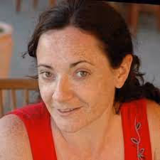 Woman with dark hair and freckles wearing a red sleeveless top, smiling softly indoors.