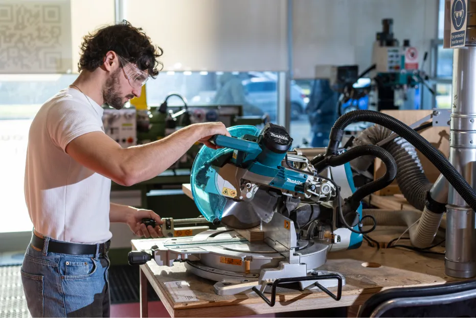 Pablo Perez wearing safety goggles using a miter saw to cut wood in a workshop.
