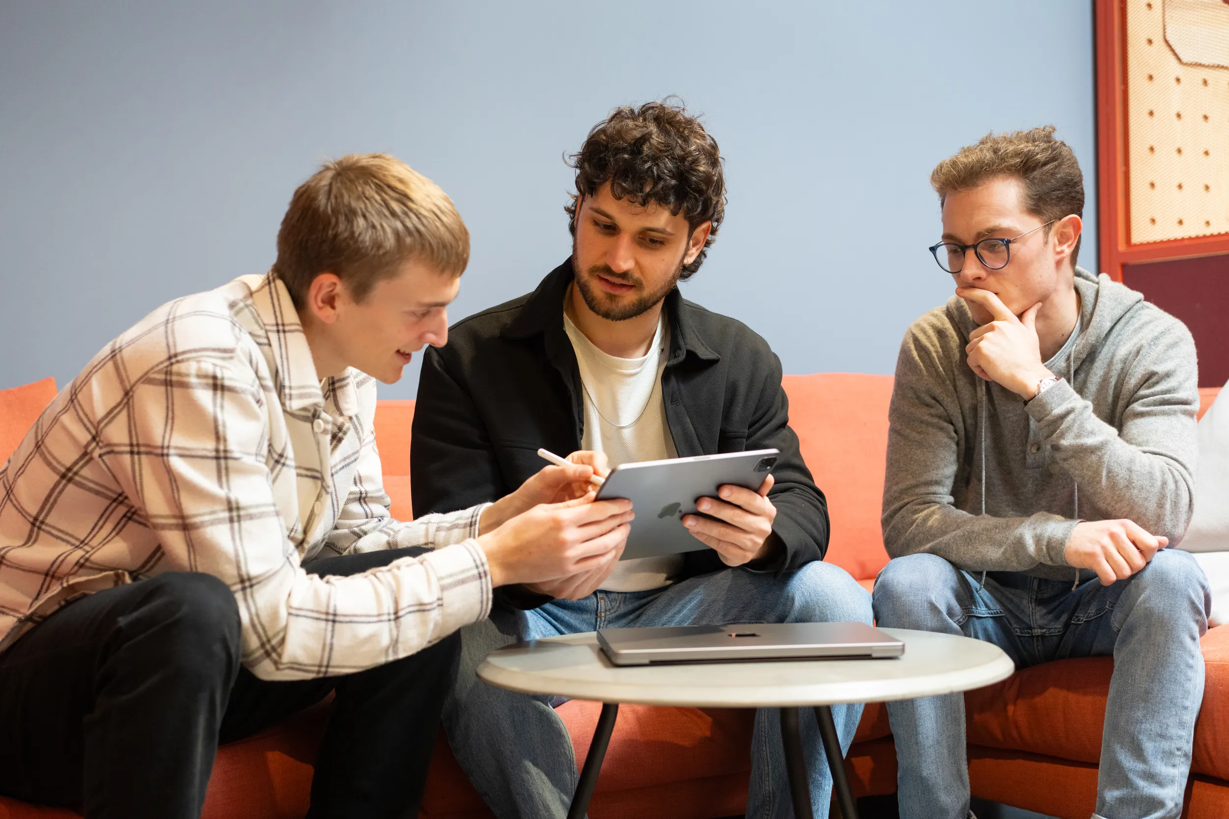 Three young men sitting on an orange couch around a round table, looking at and discussing content on a tablet.