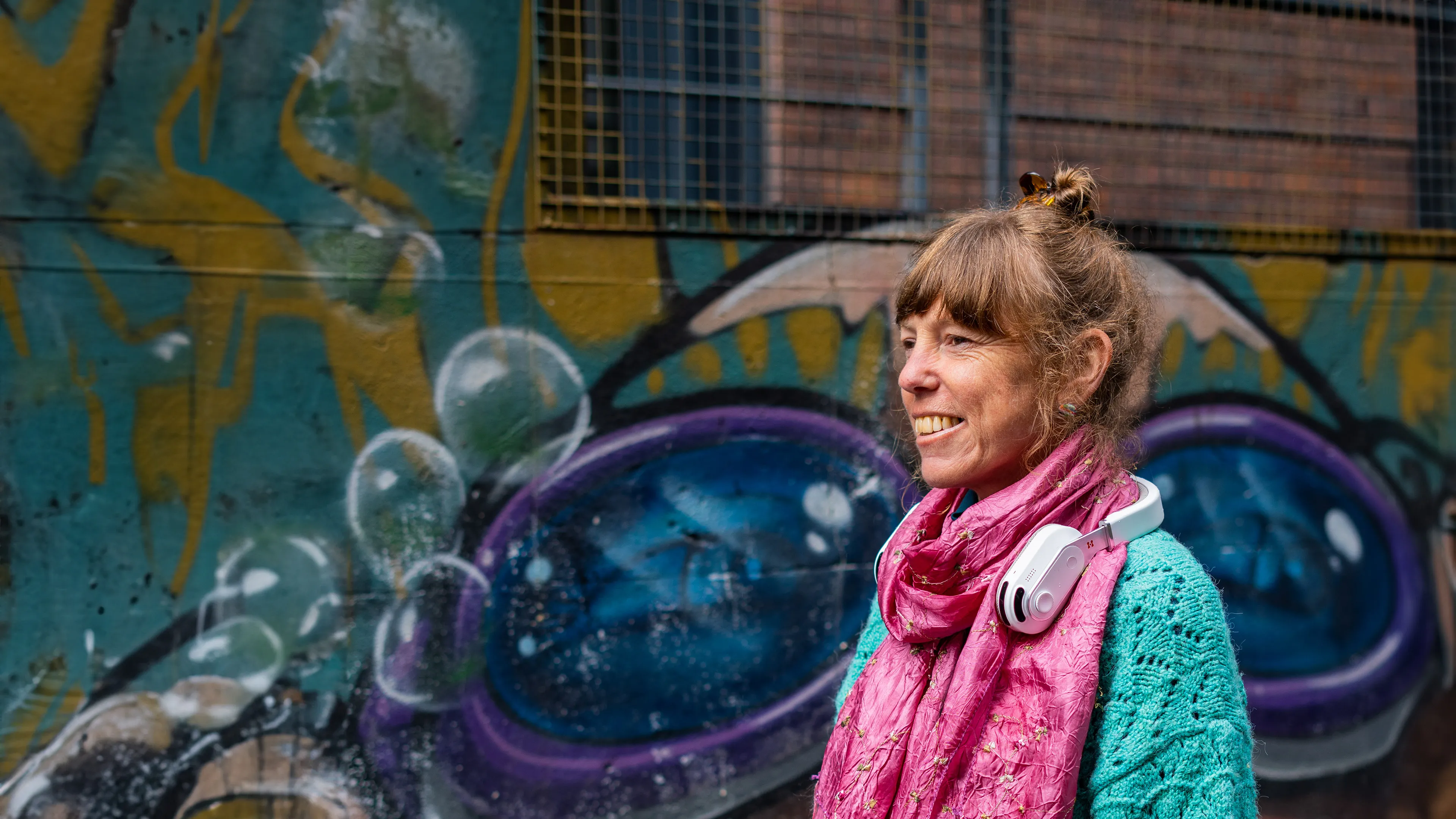 Smiling woman wearing the Sixth Sense product by Hope Tech around her neck, standing in front of a graffiti wall with colorful abstract art.