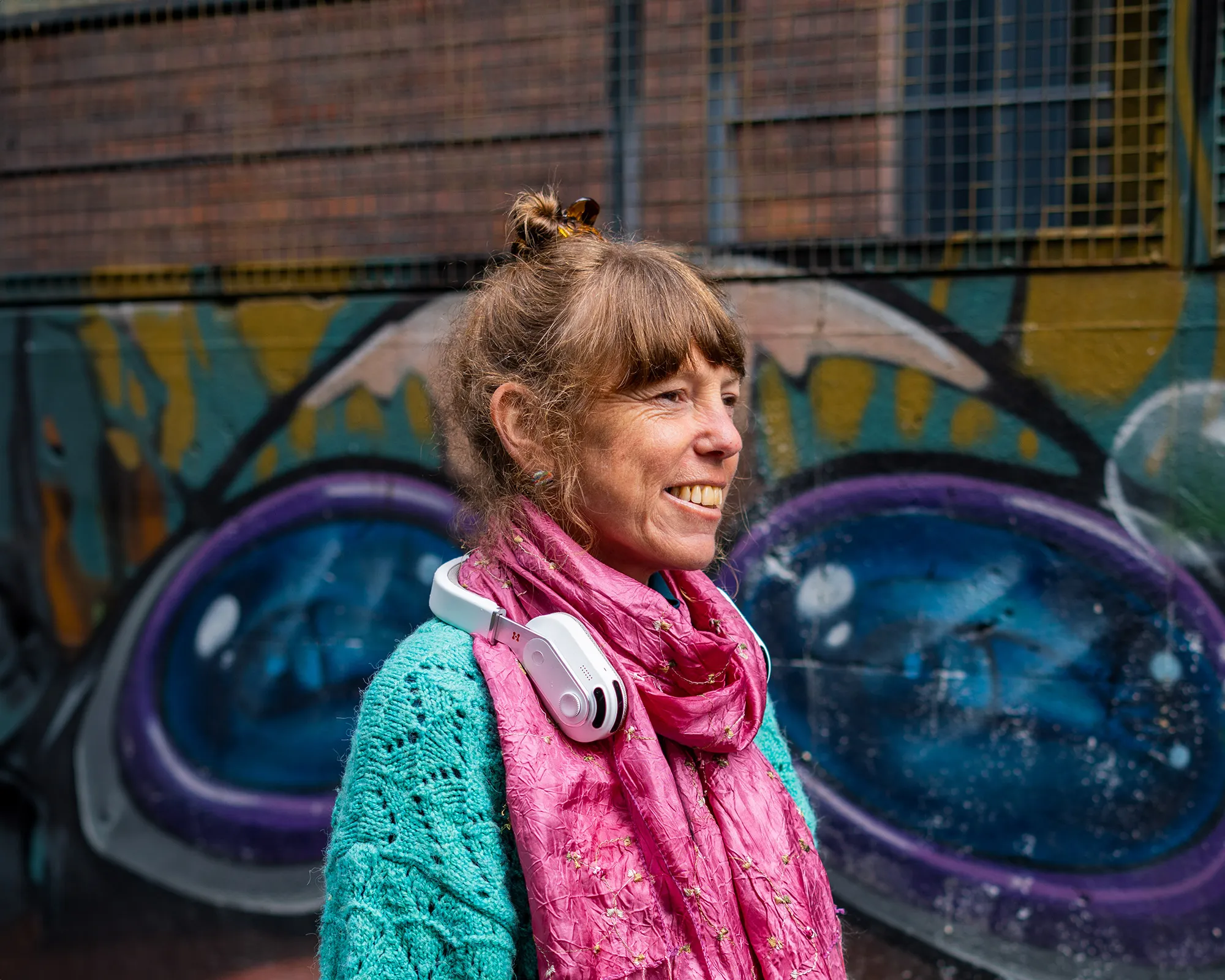 Smiling woman wearing the Sixth Sense product by Hope Tech around her neck, standing in front of a graffiti wall with colorful abstract art.