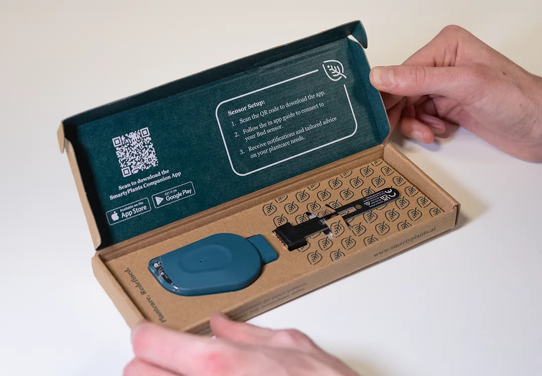 Person opening a box containing a green SmartyPlants sensor device with setup instructions and a QR code inside the lid.