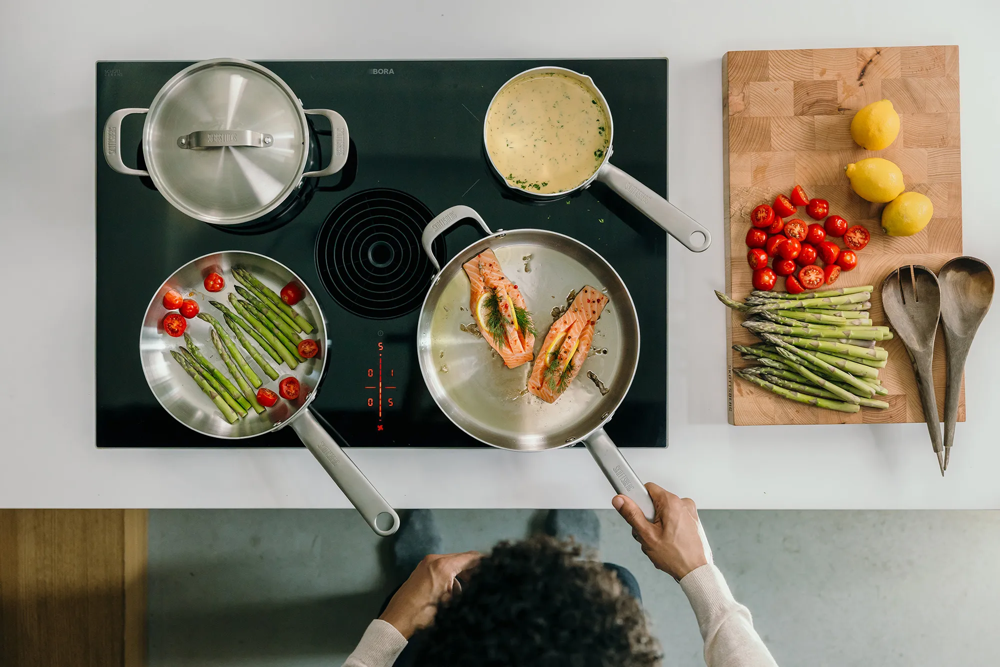 Top-down view of a person cooking salmon in a pan on an induction stove with asparagus and cherry tomatoes in another pan, a pot of sauce, and a cutting board with lemons, asparagus, cherry tomatoes, and wooden utensils.