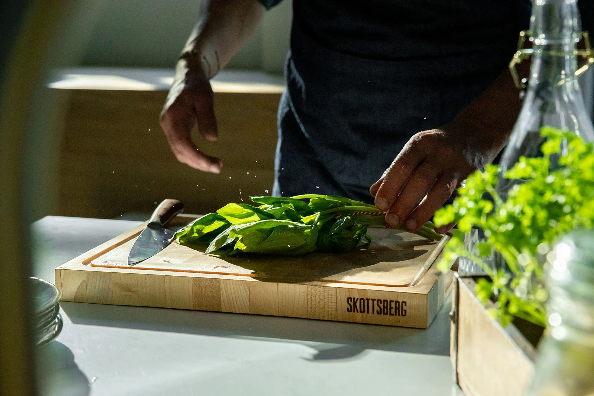 Person's hands holding fresh green leafy vegetables on a wooden Skottsberg cutting board with a knife nearby.