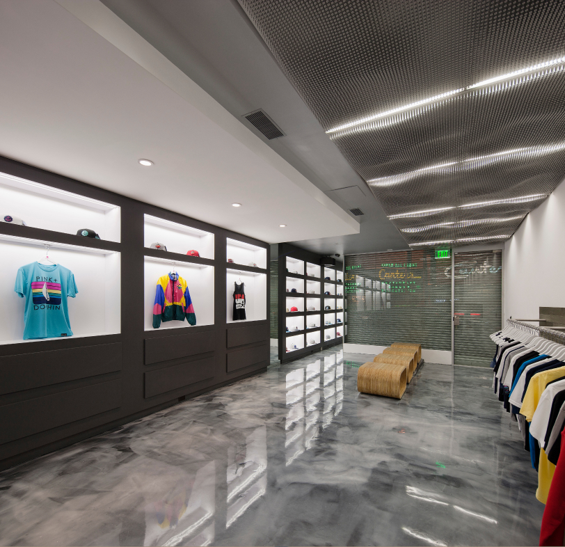 Modern retail store interior with polished concrete floor, illuminated display shelves holding clothing and hats, a bench in the center, and clothing racks on the right wall.