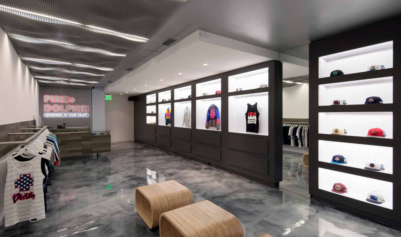 Modern retail store interior with illuminated shelves displaying apparel and caps, polished gray marble floor, and a neon sign reading 'PINK+DOLPHIN LEGENDS AT OUR CRAFT.'