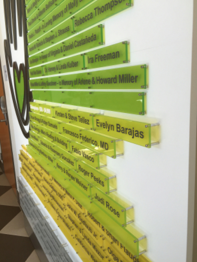 Wall display with multiple translucent green and yellow rectangular plaques featuring engraved names and dedications, mounted in a staggered pattern.