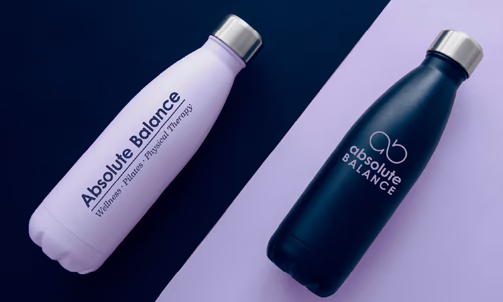 Two, high contrast, water bottle are angled in the image, with a split screen colored background. A lilac bottle on a navy background shows one logo for Absolute Balance • Wellness • Pilates • Physical Therapy. Next to it, a navy bottle on a lilac background shows a different logo for the same business.