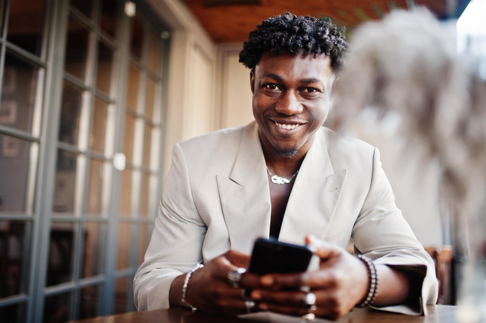Smiling man in white blazer holding a smartphone and sitting at a wooden table indoors.