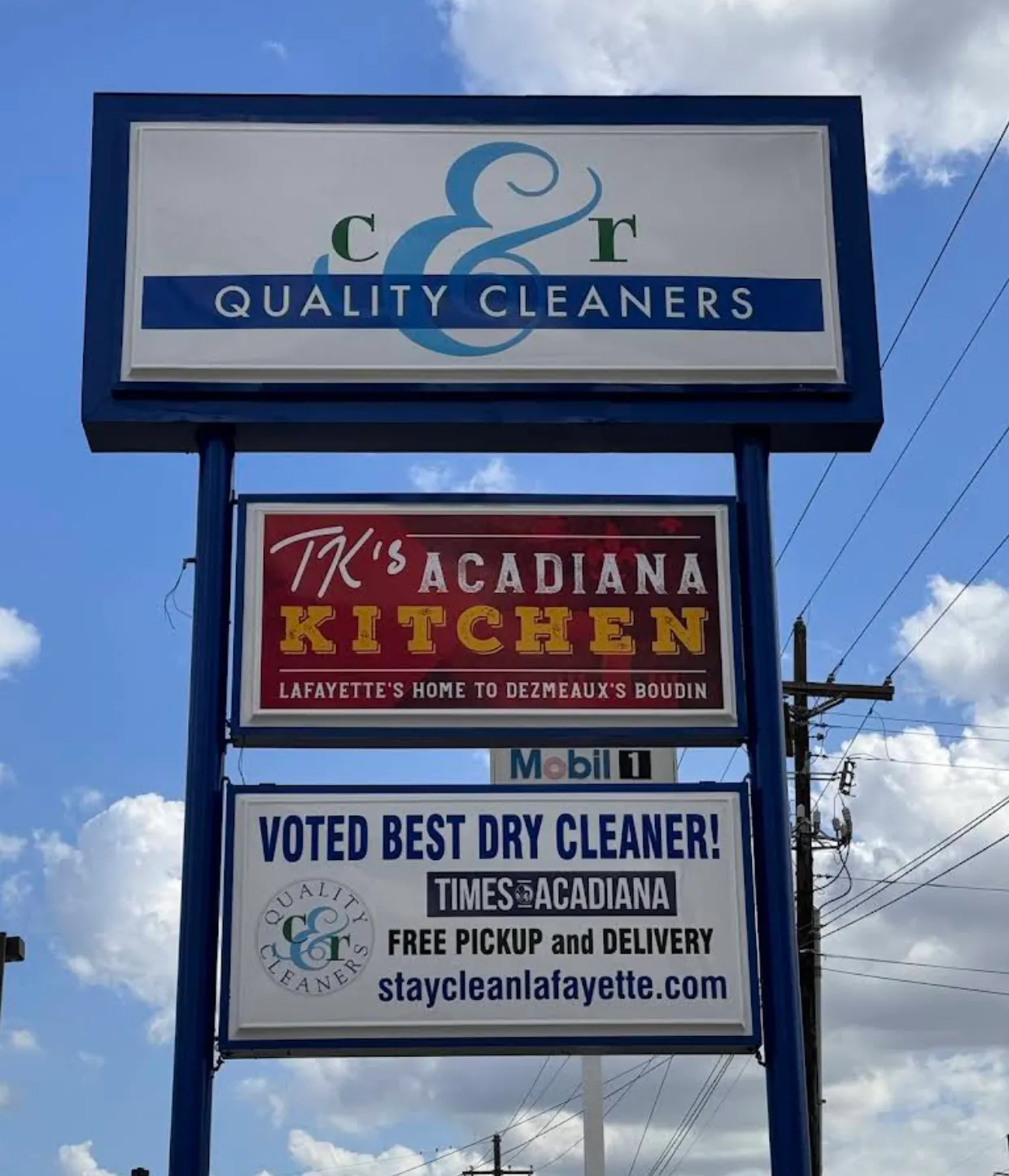 Signpost with C&R Quality Cleaners and TK's Acadiana Kitchen advertisements under a blue sky.