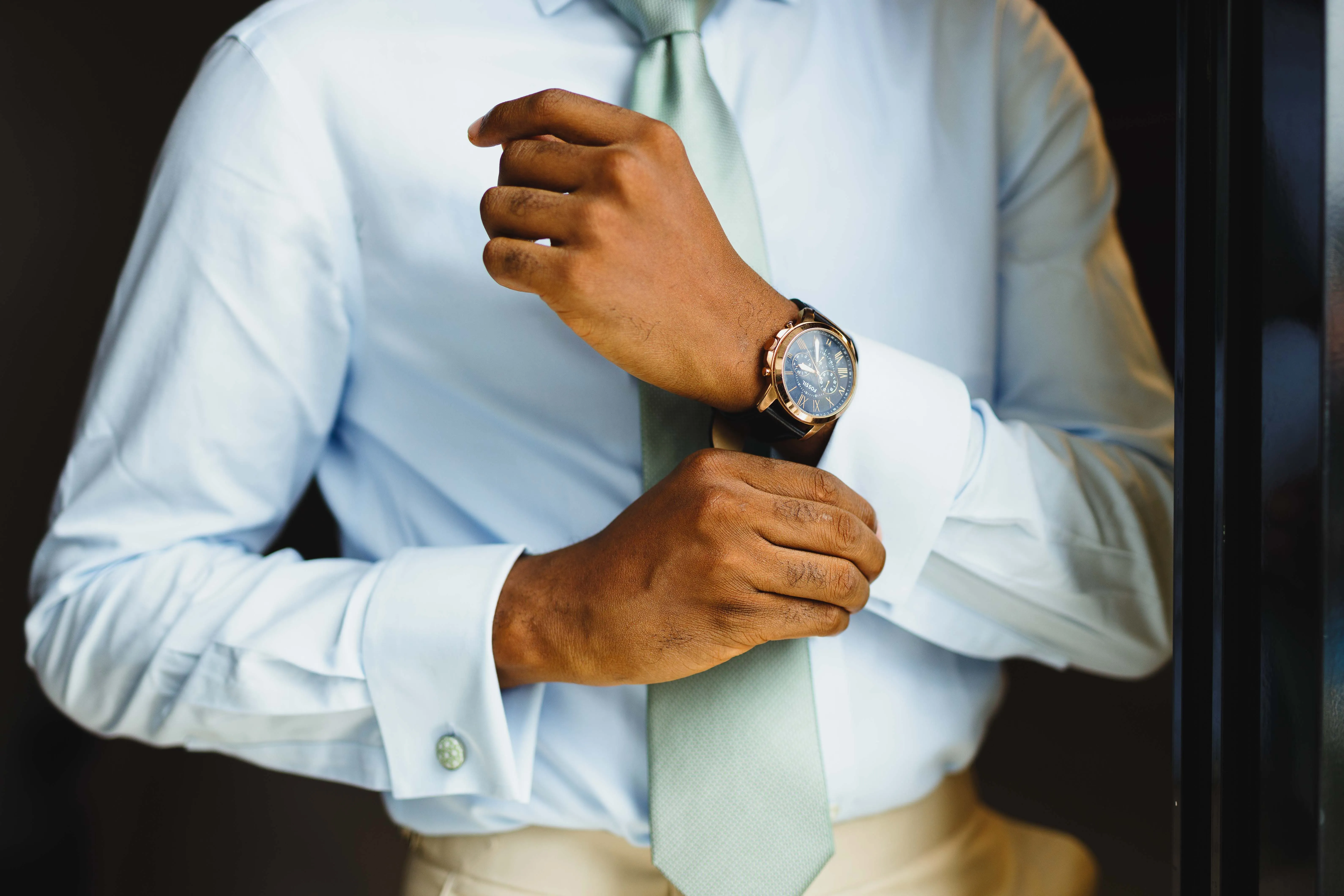 Man wearing light blue dress shirt adjusting cuff with wristwatch on left wrist.