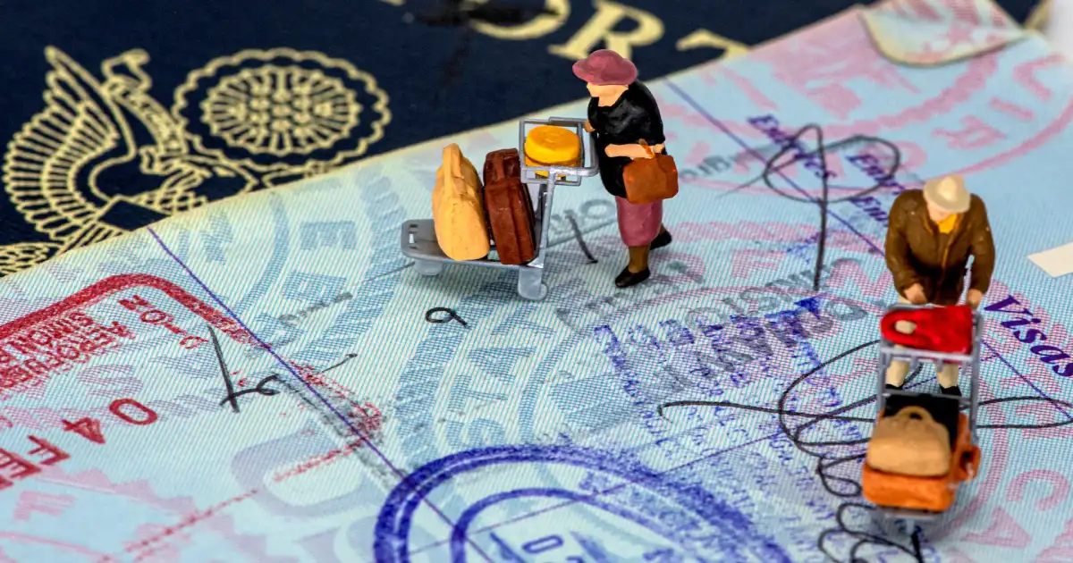 Fotografia em detalhe de uma folha de passaporte carimbada com um visto com duas miniaturas de viajantes andando sobre o passaporte carregando malas e carrinhos de aeroporto