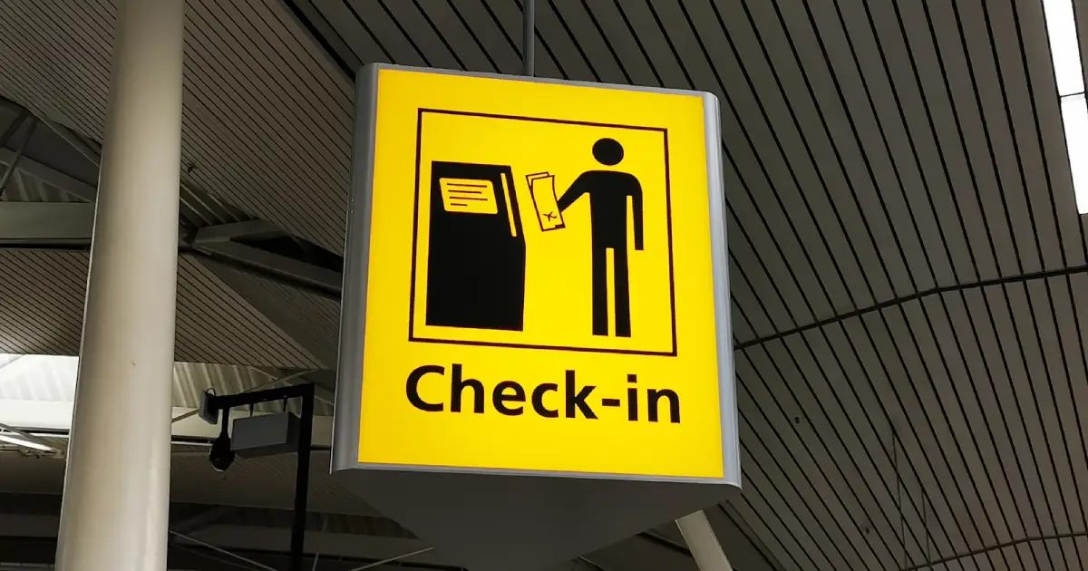 Fotografia de uma placa de aeroporto onde lê-se "CHECK-IN" em preto sobre fundo amarelo com um desenho estilizado de uma pessoa acessando uma máquina de autoatendimento para check-in