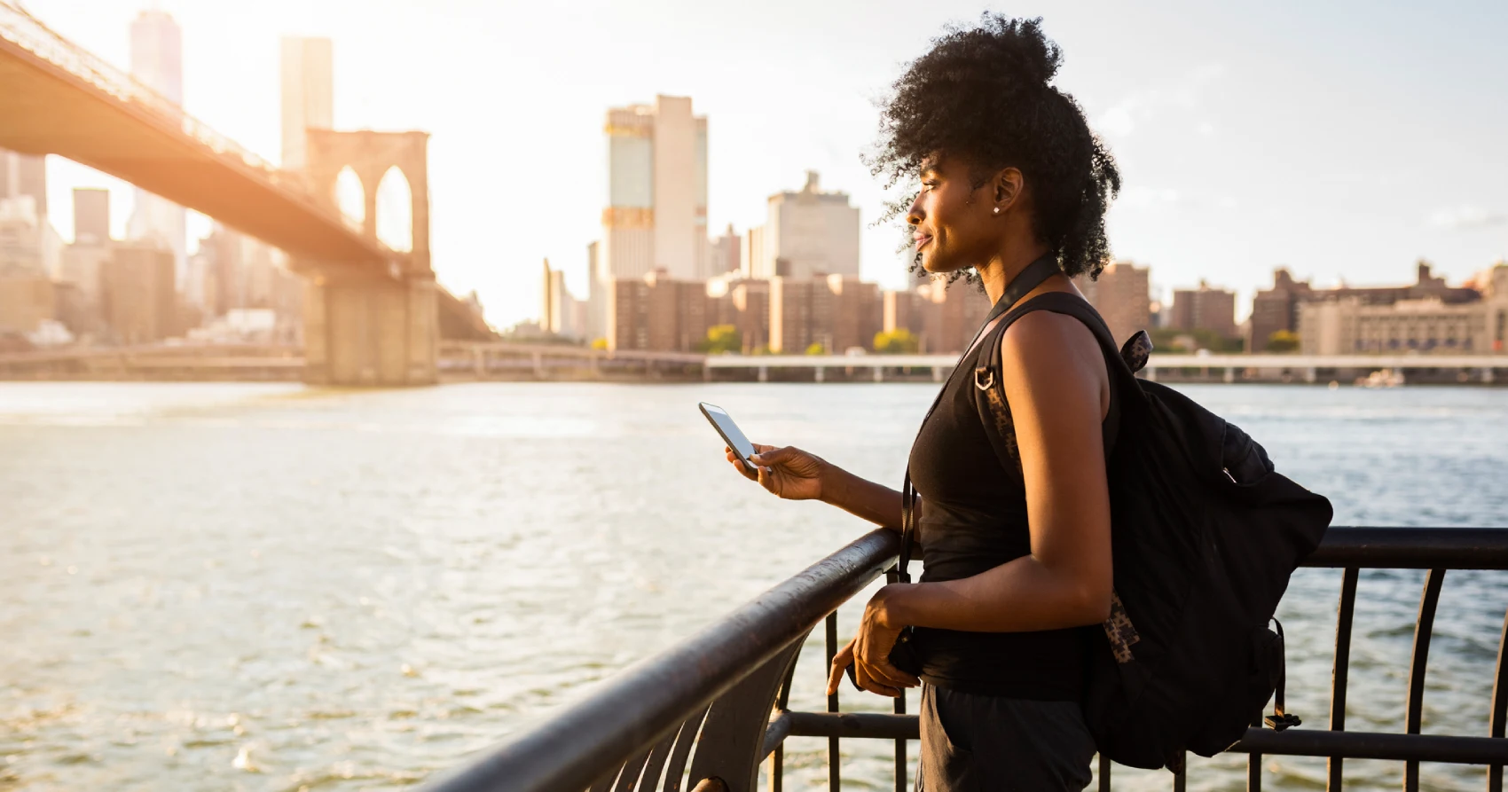 Mulher negra com celular na mão parada em frente a um guarda-corpo olhando para uma paisagem urbana durante o pôr-do-sol