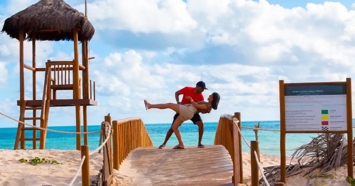 Fotografia de um casal em uma praia posicionados no fim de uma passarela de madeira, no centro da imagem. O homem, vestido com camisa laranja e bermuda preta, carrega a mulher, vestida com camisa verde-musgo e short bege claro. Do lado direito da imagem, uma placa de avisos. Do lado esquerdo, uma torre de vigia de salva-vidas.