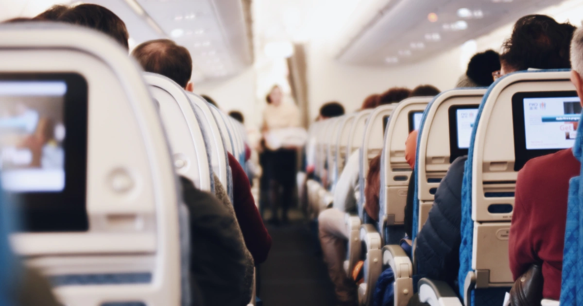 Fotografia colorida de um corredor de avião mostrando, de ambos os lados, poltronas ocupadas com passageiros. Ao fundo, em desfoque, uma comissária de bordo em pé.