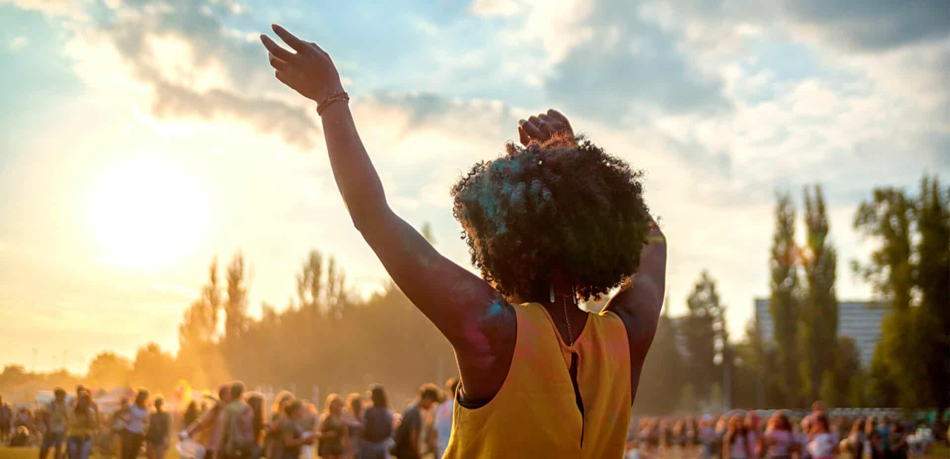 Fotografia de uma mulher negra de costas dançando em um festival. Ao fundo, imagem desfocada de pessoas dançando e árvores compondo a paisagem.