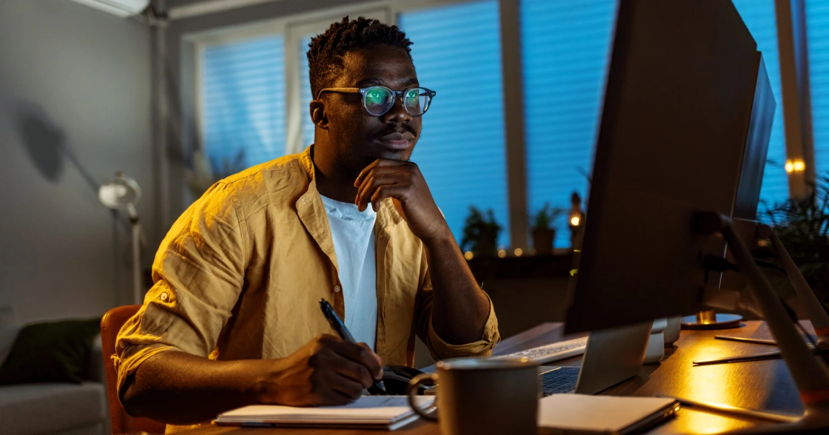 Homem jovem sentado à uma escrivaninha usando um computador e fazendo anotações em um caderno