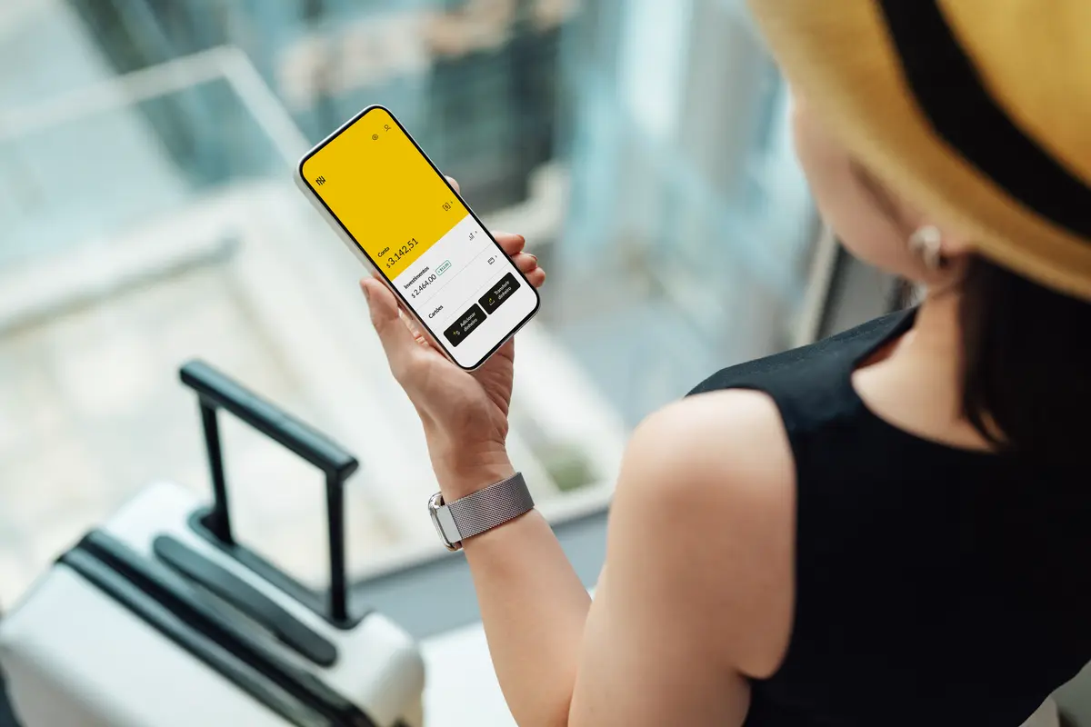 Mulher segurando celular com aplicativo da Nomad e uma mala no aeroporto