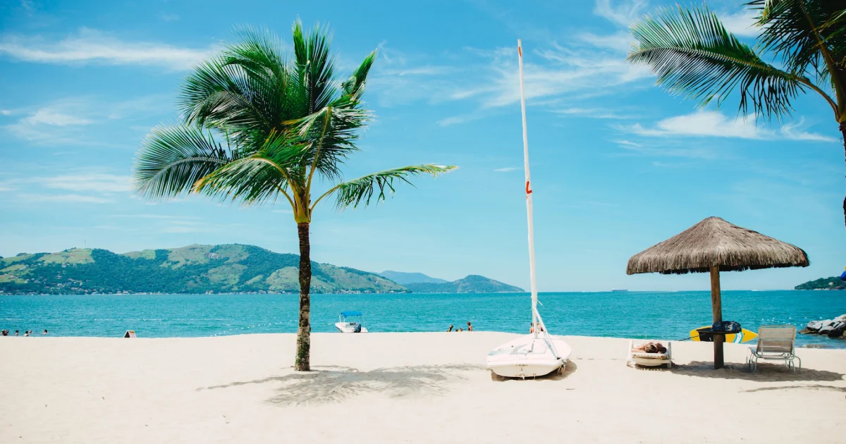 Imagem de uma praia com areia branca, palmeiras e um quiosque