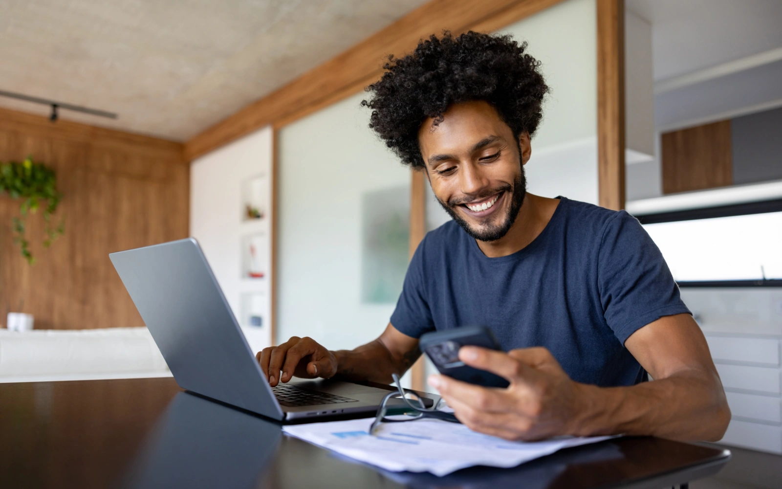 Homem de camisa azul usando celular e digitando em notebook