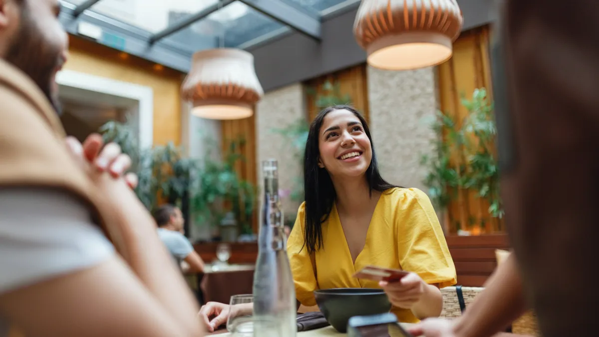 Mulher jovem sorridente, vestindo uma blusa amarela, em um restaurante com iluminação natural. Ela segura um cartão de débito internacional para pagar a conta em uma maquininha, representando a facilidade de uso da conta Nomad em viagens internacionais.