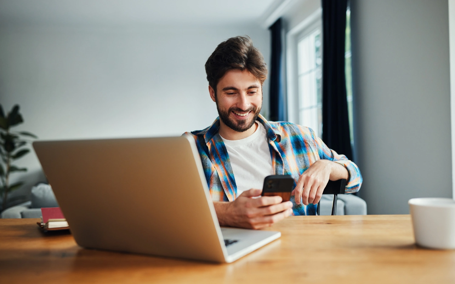 Homem de camiseta branca e camisa xadrez sentado à escrivaninha com notebook e celular