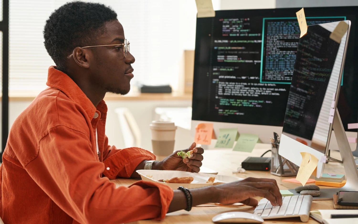 Homem negro de óculos com jaqueta laranja usando computador com duas telas para programar