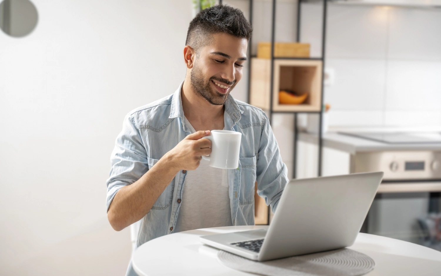 Homem branco de camiseta branca e camisa cinza usando notebook e bebendo de uma xícara