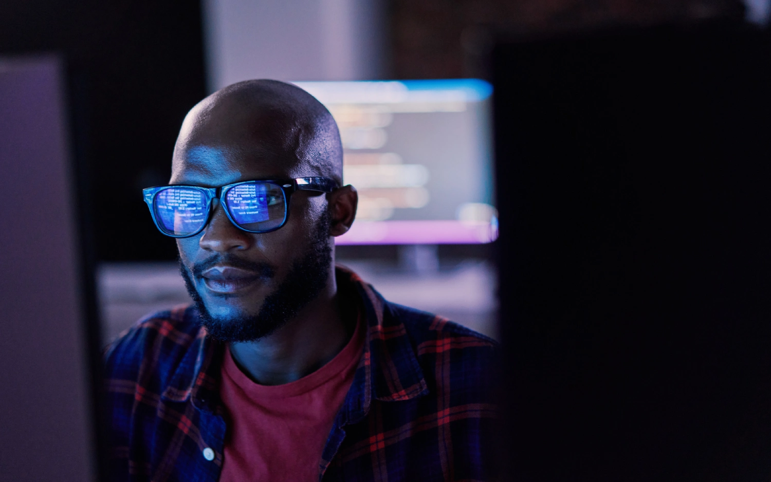 Homem negro usando óculos de aro grosso sentado em frente à monitor de computador em um ambiente escuro