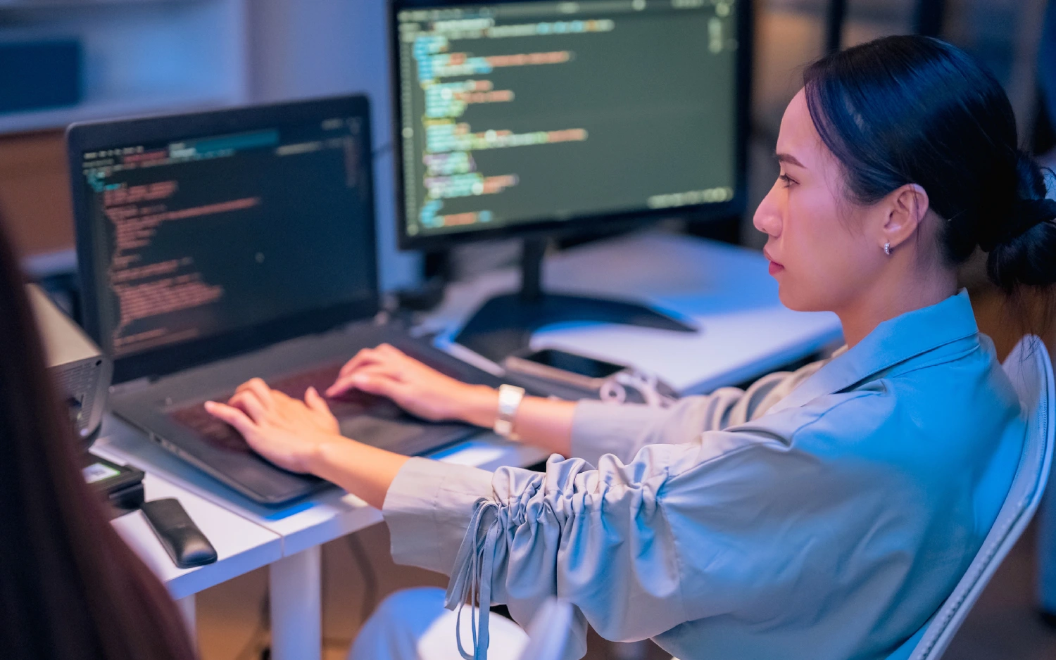 Mulher de camisa de manga longa azul usando computador com duas telas para programar