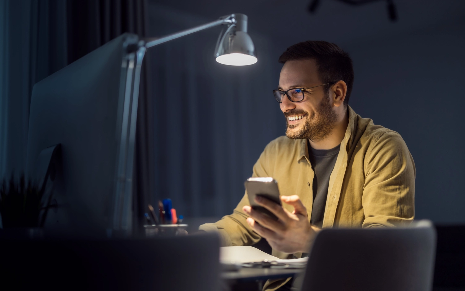 Homem de óculos e camisa amarela sorrindo enquanto usa um celular e olha para a tela de um computador