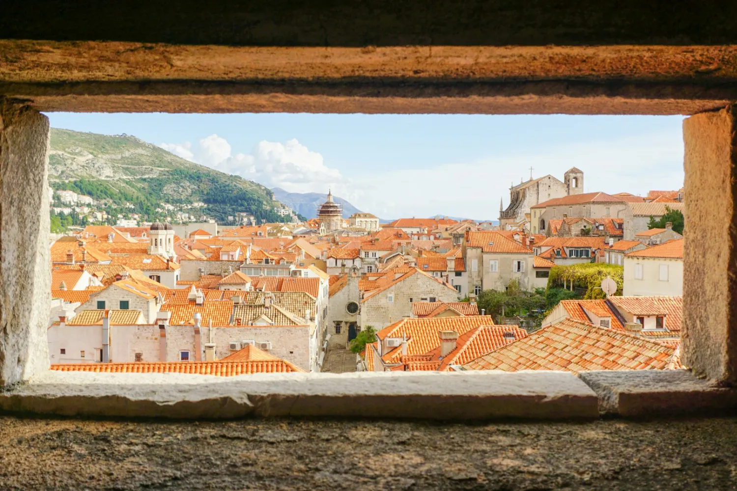 Paisagem da Cidade Antiga de Dubrovnik, com igrejas, construções antigas e montanhas ao fundo, vista através de uma janela