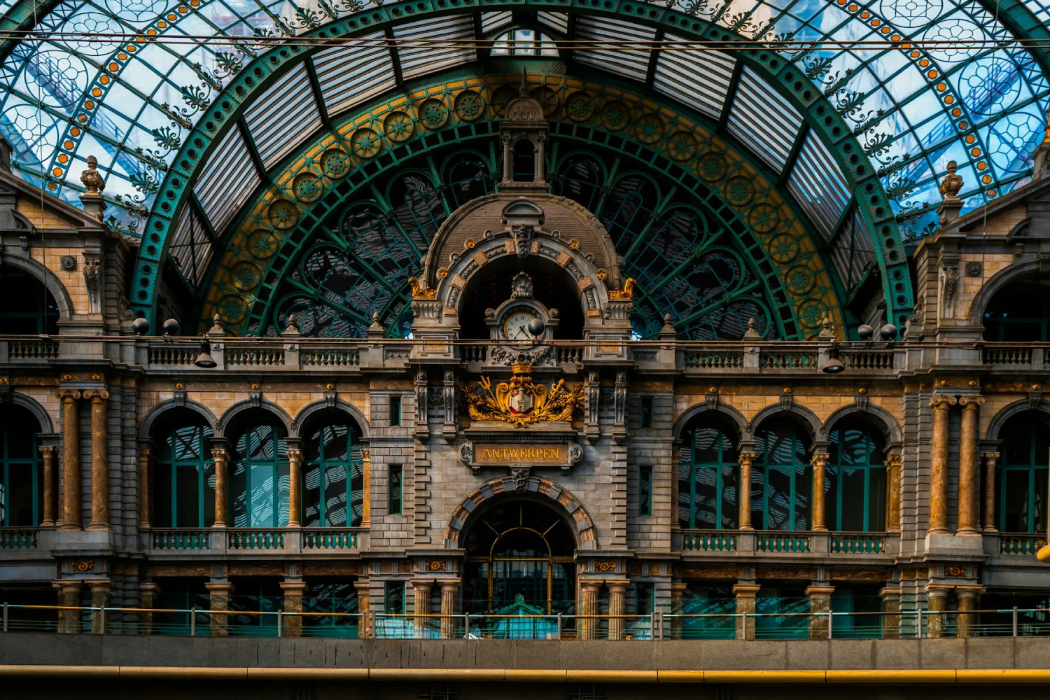 Visão interna da Estação Central de Antuérpia, com teto de vidro ornamentado em ferro e um grande relógio central