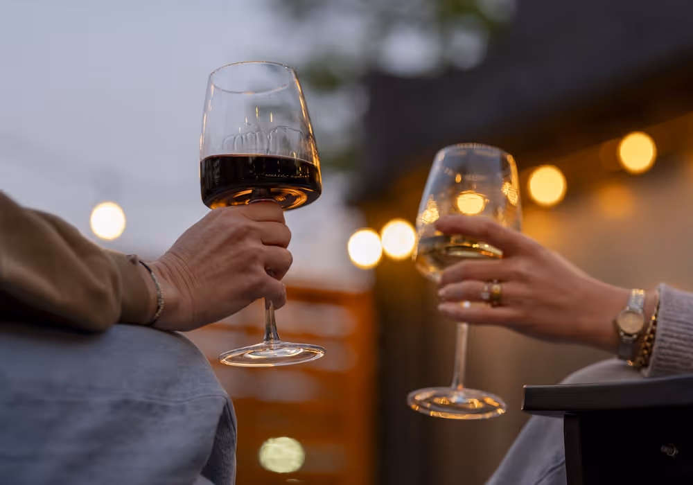 Two people clinking wine glasses outdoors in the evening, one holding red wine and the other white, with warm string lights glowing softly in the blurred background.